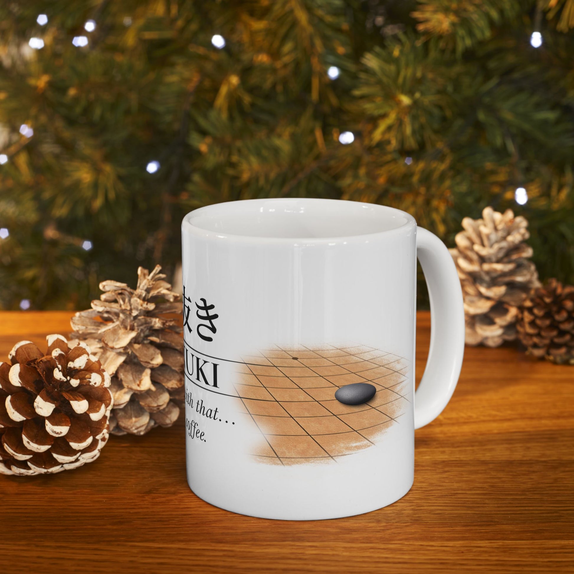 White mug with a game of Go design on a wooden surface with pine cones and a Christmas tree in the background.
