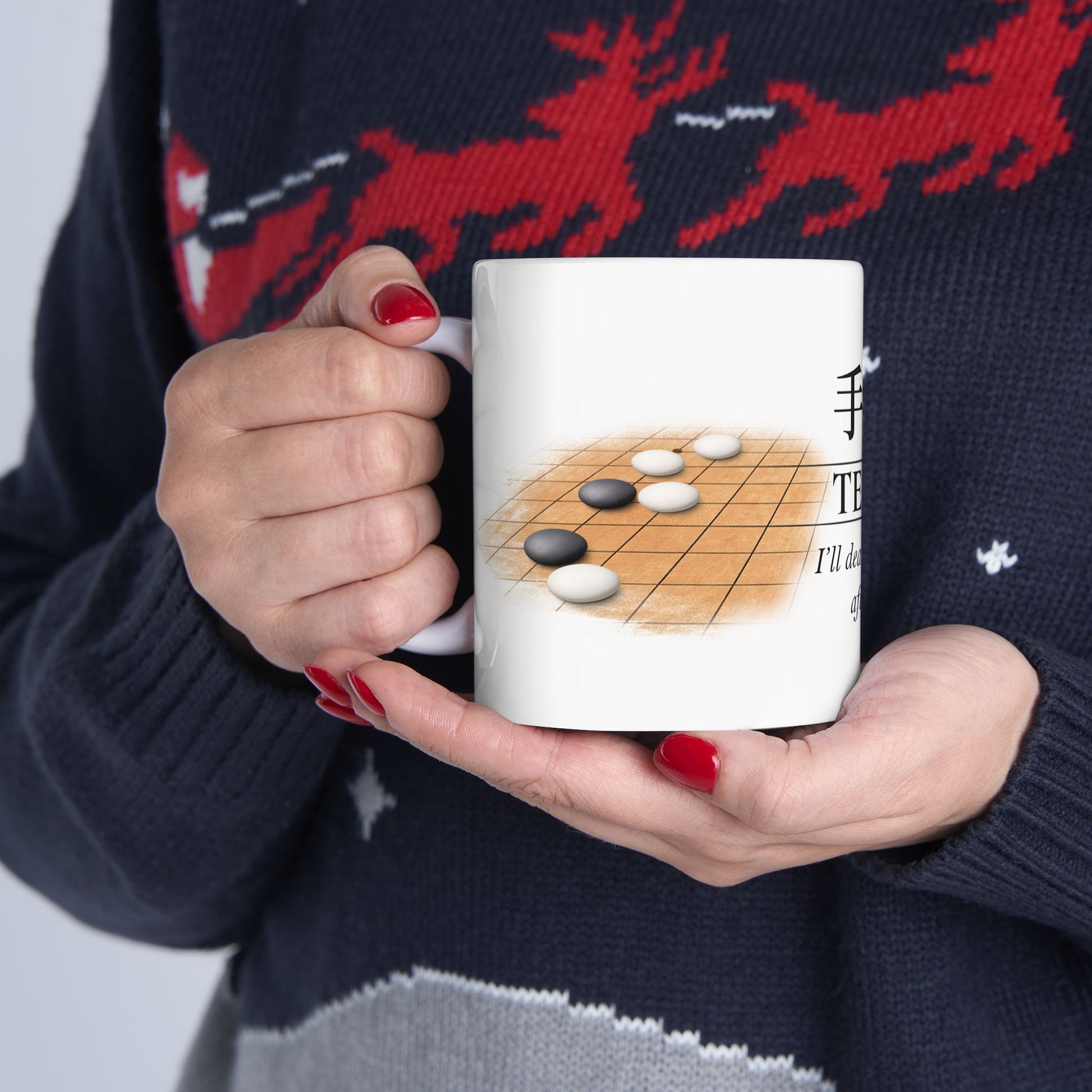 Person holding a mug with a game of Go design, wearing a dark sweater with red deer patterns.