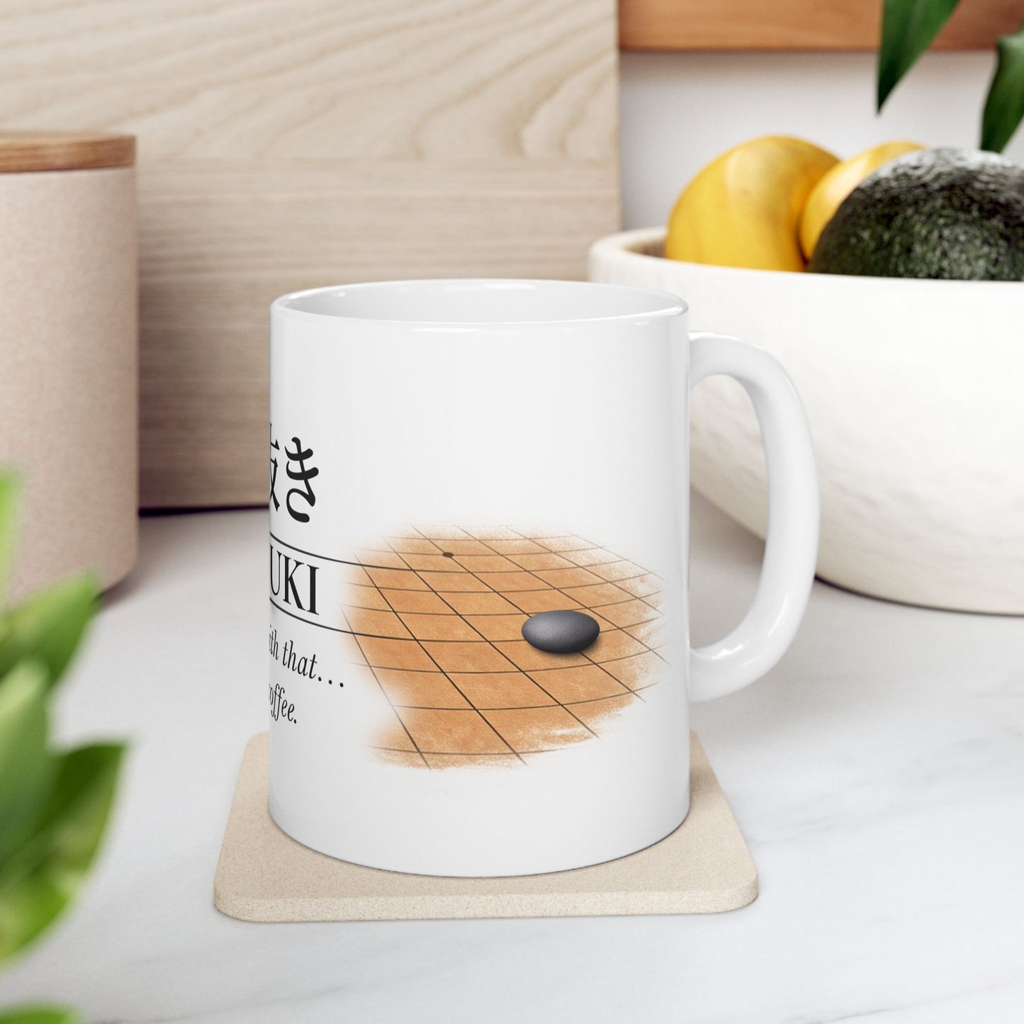 White mug with a design of a stone on a grid, placed on a coaster with a wooden surface and fruits in the background.